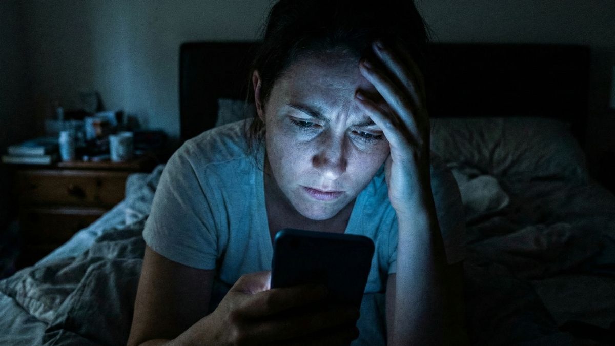 Una mujer con expresión de agobio y preocupación mira la pantalla de su teléfono móvil en una habitación a oscuras por la noche.